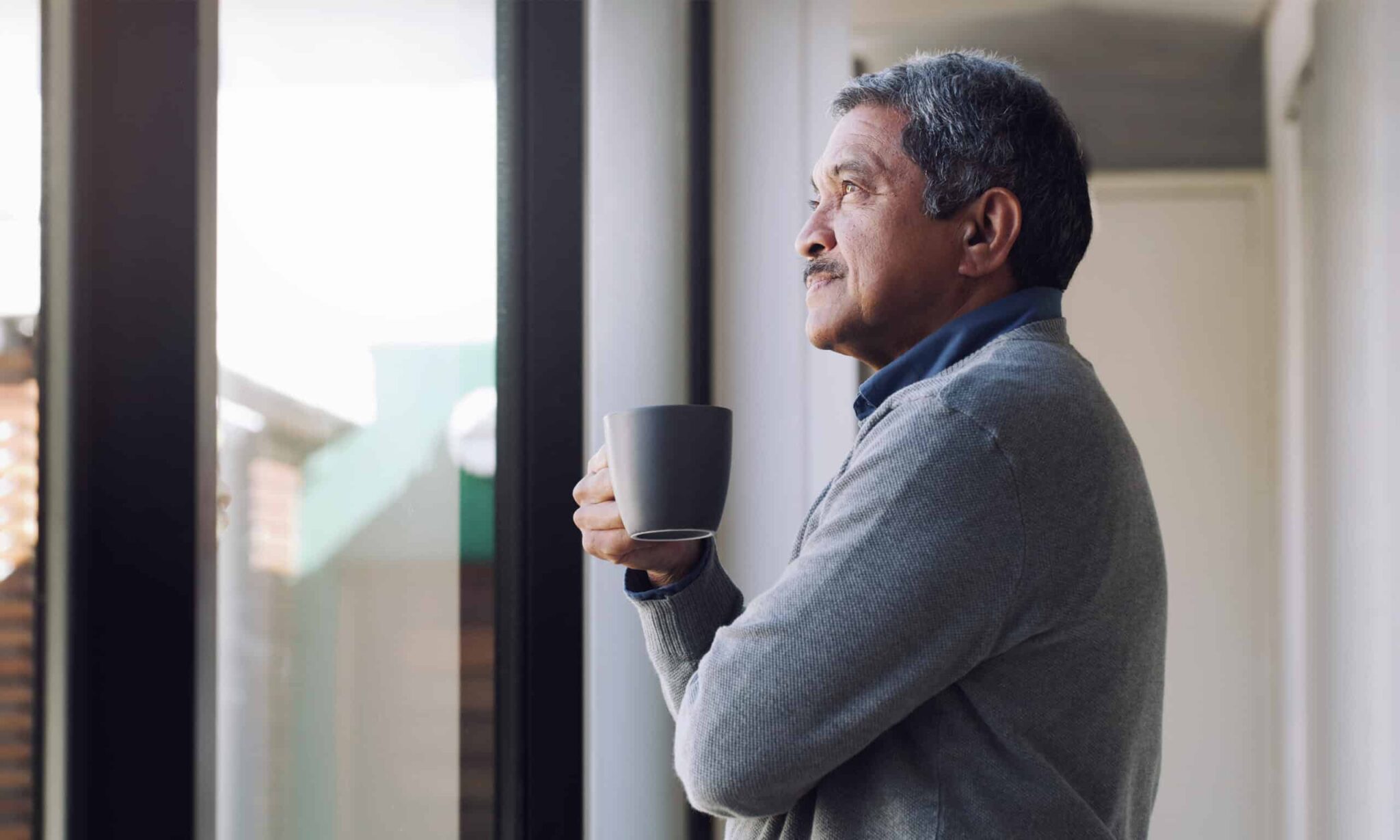 Older man drinking coffee and looking thoughtfully out his window.