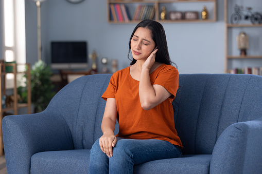 Woman in an orange shirt sitting on a couch, holding her ear experiencing ear pain and hearing loss.